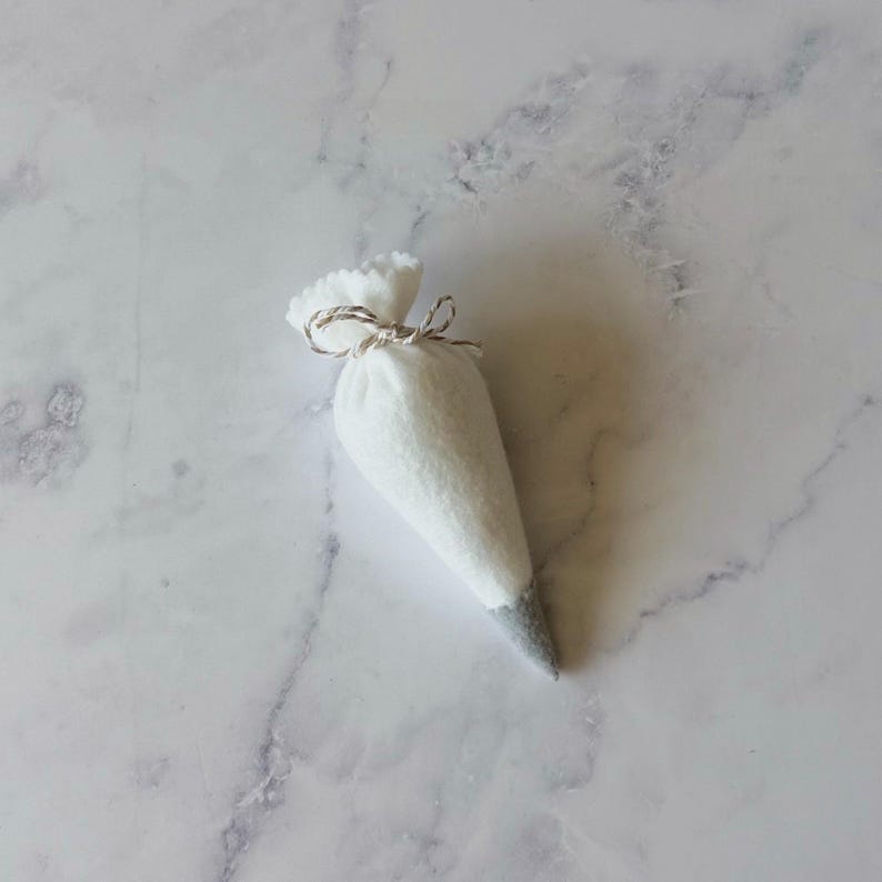 Puede incluir: Una manga pastelera de fieltro blanco con una punta gris, atada en la parte superior con un cordel fino de color natural. El objeto en forma de cono presenta una construcci&oacute;n de tela suave y texturizada con un dise&ntilde;o simple y funcional.