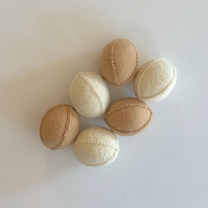 Puede incluir: Un conjunto de seis bolas de lana fieltrada de forma ovalada dispuestas en grupo. El set incluye tres bolas de color beige claro y tres bolas de color crema blanquecino, cada una con una costura visible que recorre el centro de la pieza.