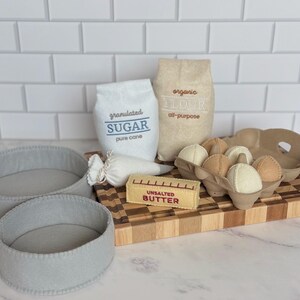 Puede incluir: Un juego de accesorios de cocina de fieltro que incluye dos moldes redondos grises, una bolsa blanca con la etiqueta granulated SUGAR, una bolsa beige con la etiqueta organic FLOUR, una manga pastelera blanca, una barra de UNSALTED BUTTER y seis huevos.