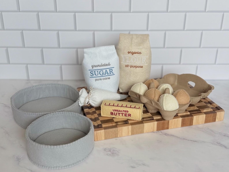 Puede incluir: Un juego de accesorios de cocina de fieltro que incluye dos moldes redondos grises, una bolsa blanca con la etiqueta granulated SUGAR, una bolsa beige con la etiqueta organic FLOUR, una manga pastelera blanca, una barra de UNSALTED BUTTER y seis huevos.