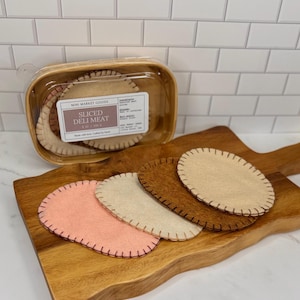 May include: A wooden cutting board displays felt deli meat slices in shades of pink, cream, and brown. A clear plastic container holds additional felt slices, labeled "SLICED DELI MEAT" with "Mini Market Goods" branding.