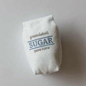 Puede incluir: Una bolsa de fieltro blanco con forma de paquete de az&uacute;car con texto bordado que dice granulated SUGAR pure cane. Las palabras granulated y pure cane est&aacute;n cosidas en gris, mientras que la palabra SUGAR est&aacute; cosida en azul con l&iacute;neas.
