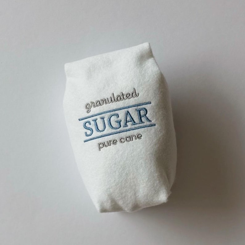 Puede incluir: Una bolsa de fieltro blanco con forma de paquete de az&uacute;car con texto bordado que dice granulated SUGAR pure cane. Las palabras granulated y pure cane est&aacute;n cosidas en gris, mientras que la palabra SUGAR est&aacute; cosida en azul con l&iacute;neas.
