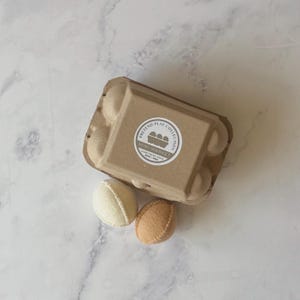 Puede incluir: Una caja de huevos de cart&oacute;n marr&oacute;n con una etiqueta circular que dice Pretend Play Collection Mini Market Farm Fresh Eggs 2019-2023. Dos huevos de fieltro, uno crema y otro tostado, est&aacute;n colocados frente a la caja con costuras visibles.