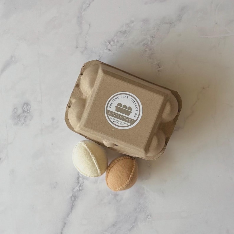 Puede incluir: Una caja de huevos de cart&oacute;n marr&oacute;n con una etiqueta circular que dice Pretend Play Collection Mini Market Farm Fresh Eggs 2019-2023. Dos huevos de fieltro, uno crema y otro tostado, est&aacute;n colocados frente a la caja con costuras visibles.