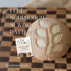 May include: A round, tan felt sourdough bread loaf featuring a decorative leaf-shaped score pattern on top. Next to the loaf is a rectangular instructional booklet titled The Mini Market Method, which displays images of sewing tools and felt food items.