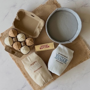 Puede incluir: Un conjunto de juguetes de fieltro que incluye una caja de cart&oacute;n con seis huevos de tela beige y crema, un cuenco de fieltro gris, una barra de fieltro rectangular con la etiqueta UNSALTED BUTTER y dos bolsas de tela con FLOUR y SUGAR.