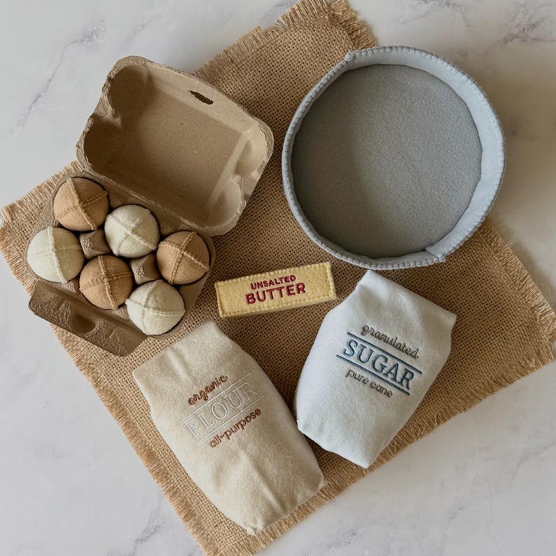 Puede incluir: Un conjunto de juguetes de fieltro que incluye una caja de cart&oacute;n con seis huevos de tela beige y crema, un cuenco de fieltro gris, una barra de fieltro rectangular con la etiqueta UNSALTED BUTTER y dos bolsas de tela con FLOUR y SUGAR.