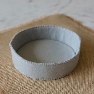 Puede incluir: Una bandeja redonda de fieltro gris claro con base plana y paredes verticales. Los bordes de la bandeja est&aacute;n unidos con puntadas de fest&oacute;n visibles y uniformes en hilo gris claro a juego, creando un recipiente circular estructurado.