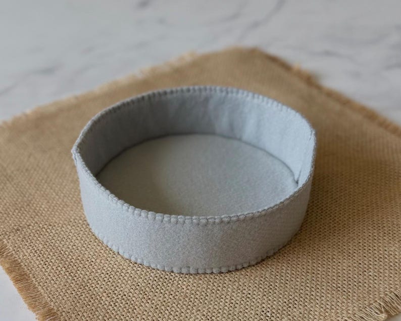Puede incluir: Una bandeja redonda de fieltro gris claro con base plana y paredes verticales. Los bordes de la bandeja est&aacute;n unidos con puntadas de fest&oacute;n visibles y uniformes en hilo gris claro a juego, creando un recipiente circular estructurado.