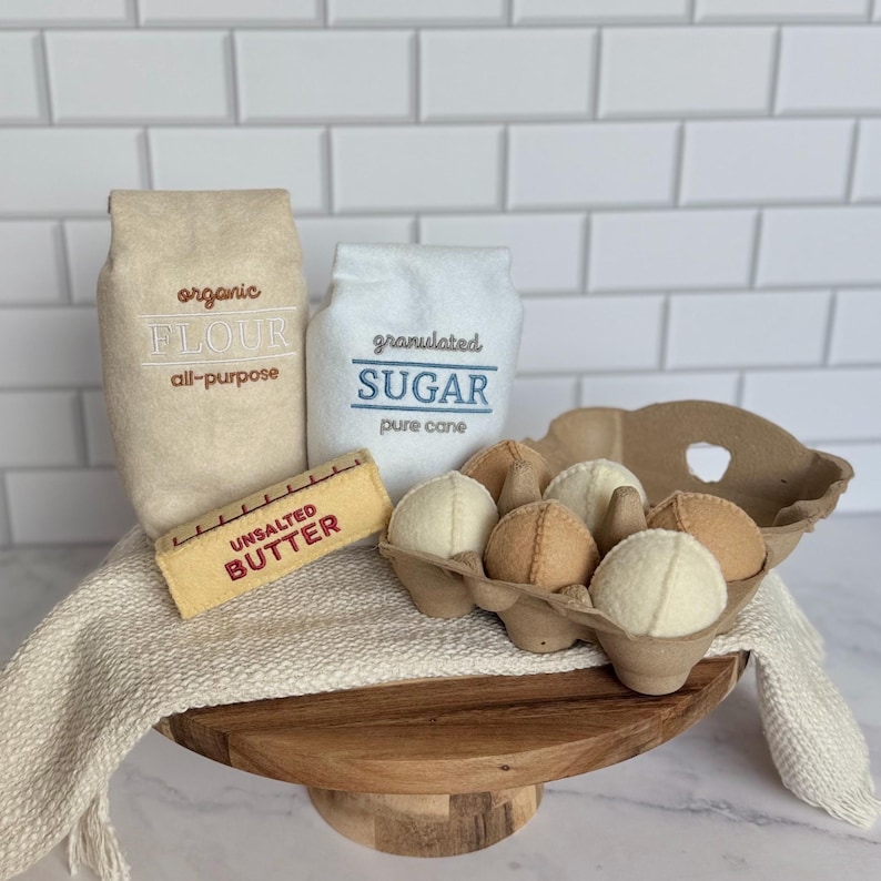 Puede incluir: Un conjunto de alimentos de fieltro que incluye una bolsa color canela etiquetada organic all-purpose FLOUR, una bolsa blanca etiquetada granulated SUGAR, una barra amarilla etiquetada UNSALTED BUTTER y una huevera de cart&oacute;n con seis huevos de fieltro.