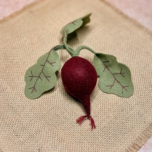 May include: A handcrafted felt radish with green leaves and red veins. The radish is a deep burgundy color, and the leaves are a light green. The radish has a small red tassel at the bottom. The radish is sitting on a tan burlap surface.