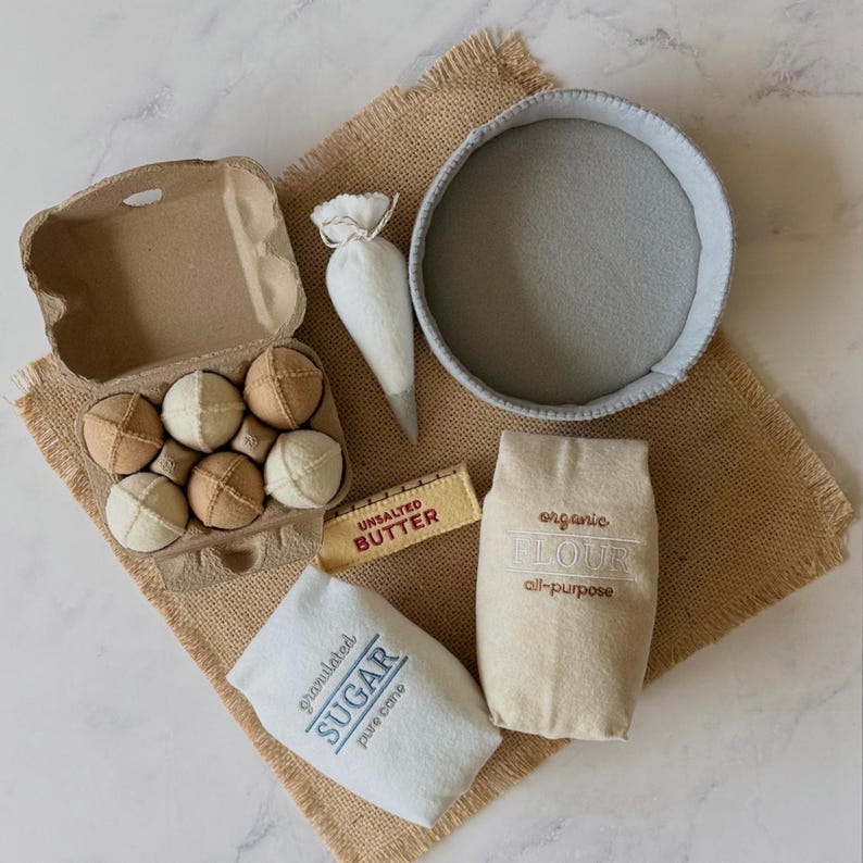 Puede incluir: Un juego de ingredientes de cocina de fieltro que incluye una caja de cart&oacute;n con seis huevos de tela, una manga pastelera blanca, un cuenco de fieltro gris, una barra de fieltro amarilla con la etiqueta UNSALTED BUTTER, una bolsa beige con organic FLOUR y una bolsa blanca con granulated SUGAR.