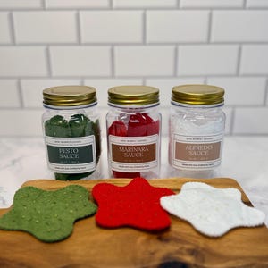 May include: Three clear plastic jars with gold lids, each containing felt shapes. Labels read Pesto Sauce, Marinara Sauce, and Alfredo Sauce. Below the jars are three star-shaped felt pieces in green, red, and white, each with small speckles.