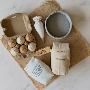 Puede incluir: Un juego de reposter&iacute;a de fieltro que incluye una huevera de cart&oacute;n con seis huevos beige y crema, una manga pastelera blanca, un cuenco gris, una barra de mantequilla amarilla, una bolsa beige con la etiqueta organic flour all-purpose y una bolsa blanca con la etiqueta granulated sugar.