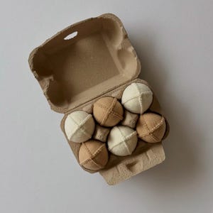 Puede incluir: Una caja de cart&oacute;n marr&oacute;n para huevos que contiene seis juguetes de tela esf&eacute;ricos. Los juguetes est&aacute;n dispuestos en dos filas, alternando entre los colores blanco crema y beige. Cada esfera de tela tiene costuras visibles en cruz.