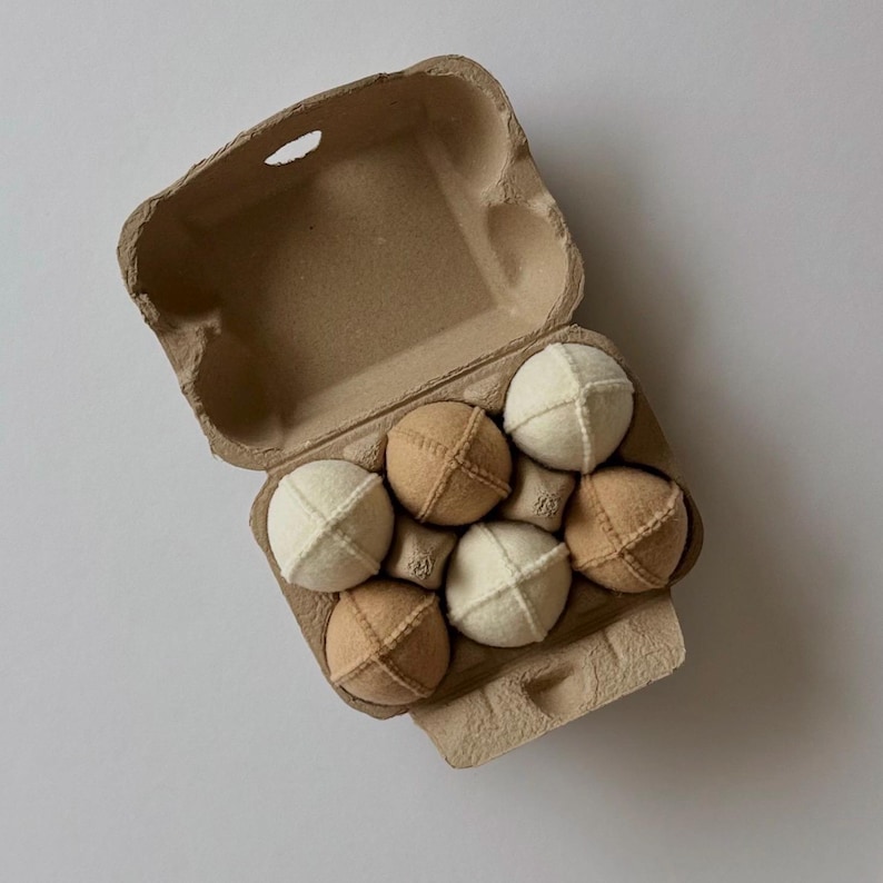 Puede incluir: Una caja de cart&oacute;n marr&oacute;n para huevos que contiene seis juguetes de tela esf&eacute;ricos. Los juguetes est&aacute;n dispuestos en dos filas, alternando entre los colores blanco crema y beige. Cada esfera de tela tiene costuras visibles en cruz.