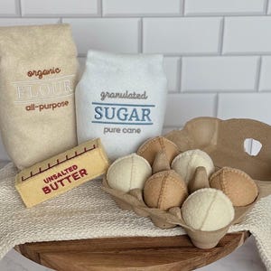 Puede incluir: Un conjunto de alimentos de fieltro que incluye una bolsa color canela etiquetada organic FLOUR all-purpose, una bolsa blanca etiquetada granulated SUGAR pure cane, una barra de mantequilla amarilla UNSALTED BUTTER y un cart&oacute;n marr&oacute;n con seis huevos de fieltro redondos.