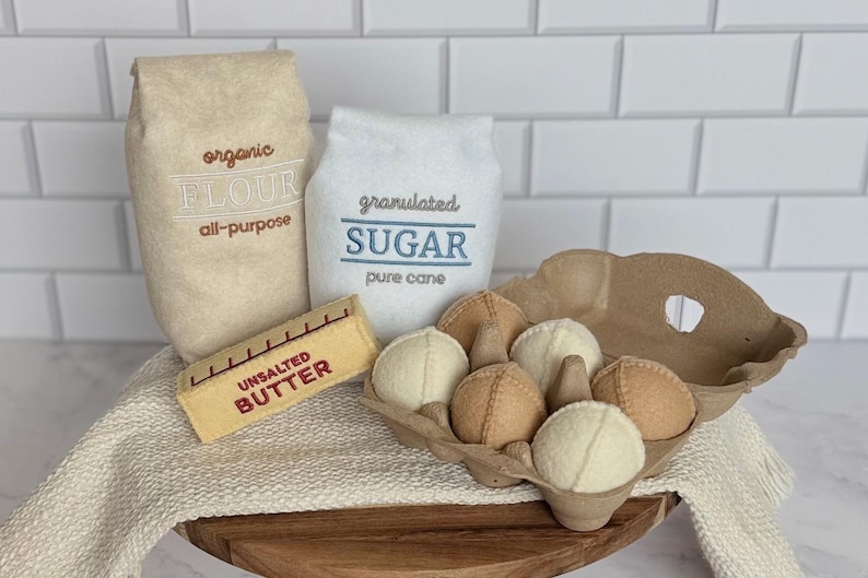 Puede incluir: Un conjunto de alimentos de fieltro que incluye una bolsa color canela etiquetada organic FLOUR all-purpose, una bolsa blanca etiquetada granulated SUGAR pure cane, una barra de mantequilla amarilla UNSALTED BUTTER y un cart&oacute;n marr&oacute;n con seis huevos de fieltro redondos.