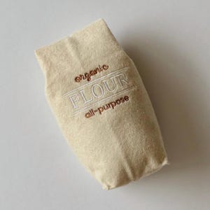 Puede incluir: Una bolsa de fieltro beige con forma de saco de harina con texto bordado que dice organic FLOUR all-purpose. Las palabras organic y all-purpose est&aacute;n cosidas con hilo marr&oacute;n, mientras que la palabra FLOUR est&aacute; cosida con hilo blanco y l&iacute;neas.