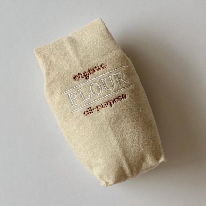Puede incluir: Una bolsa de fieltro beige con forma de saco de harina con texto bordado que dice organic FLOUR all-purpose. Las palabras organic y all-purpose est&aacute;n cosidas con hilo marr&oacute;n, mientras que la palabra FLOUR est&aacute; cosida con hilo blanco y l&iacute;neas.