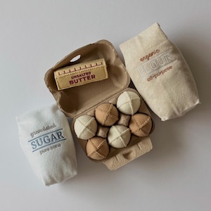 Puede incluir: Un conjunto de juguetes de tela que incluye una caja de cart&oacute;n marr&oacute;n con seis huevos redondos de tela en tonos beige y tostado, una barra de mantequilla sin sal de tela beige, una bolsa de tela blanca con la etiqueta granulated SUGAR y una bolsa de tela beige con la etiqueta organic FLOUR.