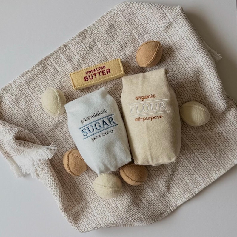 Puede incluir: Un conjunto de alimentos de juguete de fieltro que incluye una barra rectangular de mantequilla sin sal, una bolsa blanca etiquetada granulated SUGAR pure cane y una bolsa beige etiquetada organic FLOUR all-purpose, junto a peque&ntilde;as bolas de fieltro.