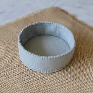 Puede incluir: Una bandeja circular de fieltro gris claro con un dise&ntilde;o poco profundo y abierto. Los bordes de la bandeja est&aacute;n rematados con una puntada de fest&oacute;n decorativa y consistente a lo largo del borde superior, dando un aspecto limpio y estructurado.