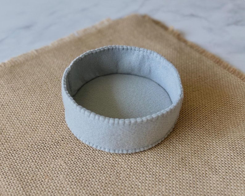 Puede incluir: Una bandeja circular de fieltro gris claro con un dise&ntilde;o poco profundo y abierto. Los bordes de la bandeja est&aacute;n rematados con una puntada de fest&oacute;n decorativa y consistente a lo largo del borde superior, dando un aspecto limpio y estructurado.