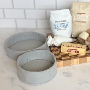 May include: A set of two grey felt round cake pans, a white felt bag labeled granulated SUGAR, a tan felt bag labeled organic FLOUR, a white felt piping bag, a yellow felt stick of UNSALTED BUTTER, and a felt egg carton with white eggs.