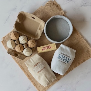 Puede incluir: Un juego de alimentos de fieltro que incluye una caja de cart&oacute;n para huevos con seis huevos de tela beige y blancos, un cuenco de fieltro gris, una barra de mantequilla de fieltro amarilla con la etiqueta UNSALTED BUTTER, una bolsa beige con la etiqueta organic FLOUR all-purpose y una bolsa blanca con la etiqueta granulated SUGAR pure cane.