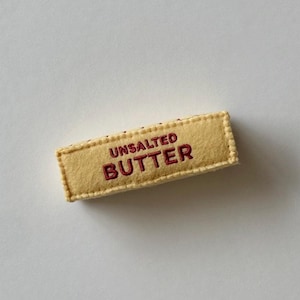 Puede incluir: Un adorno rectangular de fieltro con forma de barra de mantequilla. El art&iacute;culo es de color amarillo p&aacute;lido con bordes cosidos y presenta el texto UNSALTED BUTTER bordado en hilo rojo oscuro en el centro de la parte frontal del objeto.