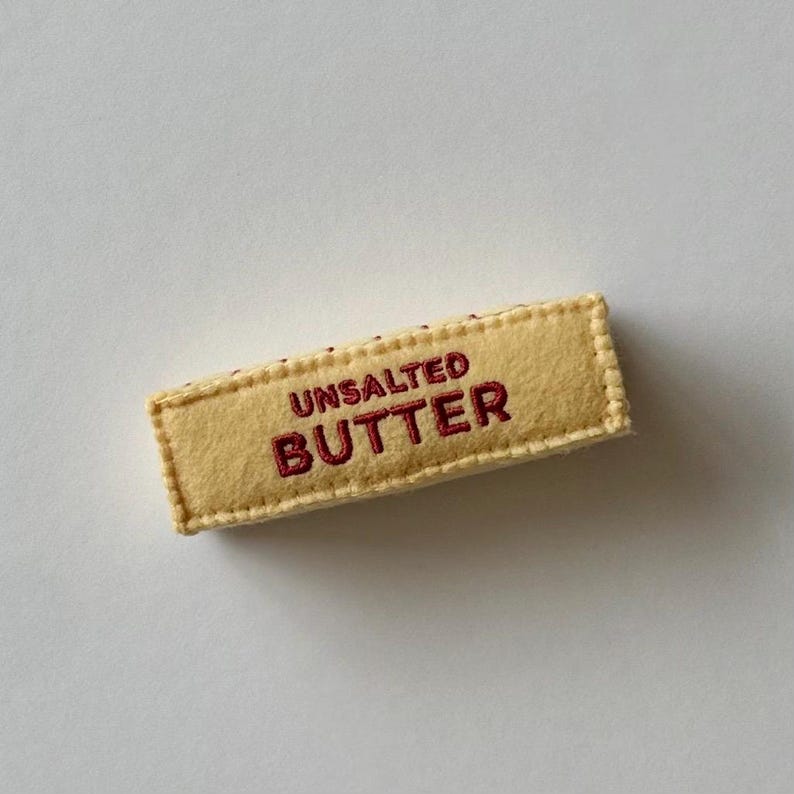 Puede incluir: Un adorno rectangular de fieltro con forma de barra de mantequilla. El art&iacute;culo es de color amarillo p&aacute;lido con bordes cosidos y presenta el texto UNSALTED BUTTER bordado en hilo rojo oscuro en el centro de la parte frontal del objeto.