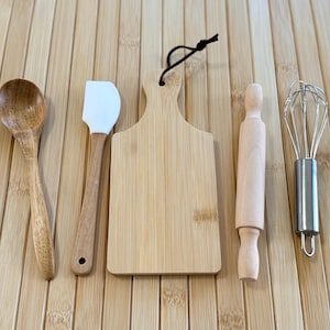 May include: A collection of wooden and stainless steel kitchen tools, including a spoon, spatula, cutting board, rolling pin, and whisk. The tools are arranged on a light-colored wooden surface.
