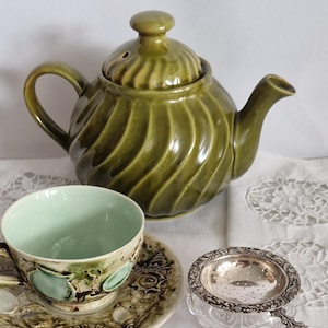 Könnte beinhalten: Eine olivgrüne Keramik-Teekanne mit Spiralmuster, eine passende Tasse und Untertasse sowie ein silberner Tee-Infuser. Tasse und Untertasse haben ein Muschelmuster. Das Innere der Tasse ist hellgrün.