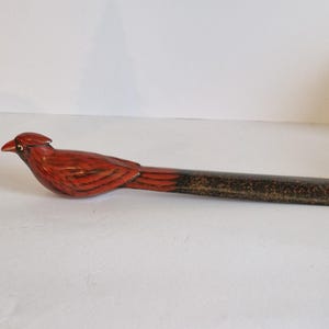 Puede incluir: Un abrecartas decorativo de madera tallada a mano con un diseño de cardenal. El pájaro está pintado en tonos rojos y marrones, con un mango largo y delgado. El abrecartas mide aproximadamente 25 cm de largo.
