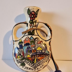 Puede incluir: Frasco de recuerdo de Jerusalén de cerámica con un diseño de paisaje urbano pintado a color. El frasco tiene una base color crema, dos asas y un cordón de cuero con una cuenta azul. La palabra "Jerusalem" está escrita en la parte delantera.