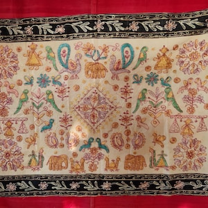Könnte beinhalten: Cremefarbener Stoff mit aufwendiger Stickerei, die Vögel, Elefanten und florale Muster in Blau-, Grün-, Gelb- und Lilatönen zeigt. Das Design wird von einem schwarzen Rand mit floralen und Blattakzenten umrahmt.