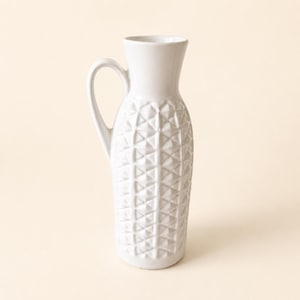 Op de afbeelding: Een witte keramische kan met een hoge, slanke hals en een enkel gebogen handvat. Het lichaam van de kan is versierd met een herhalend geometrisch patroon van verhoogde driehoeken, wat zorgt voor een getextureerd oppervlak over het onderste deel.