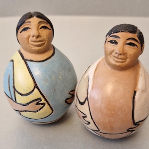 Puede incluir: Dos figuras de cerámica con caras sonrientes. Una está pintada de azul claro y amarillo, la otra de color melocotón y blanco. Ambas tienen contornos negros y tienen forma de huevo, lo que sugiere un estilo de arte popular.