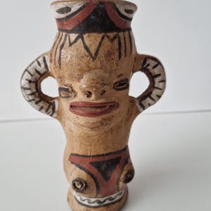 May include: A tan ceramic vessel shaped like a human figure with two curved handles. The piece features painted black and red geometric patterns on the rim, face, and torso, with a wide mouth, prominent lips, and a textured, weathered surface finish.