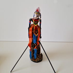 Puede incluir: Títere tradicional indonesio Wayang Golek. El títere viste un traje rojo, naranja y azul con detalles intrincados. Tiene una cara pintada y está montado sobre un soporte negro con dos varillas negras para la manipulación.