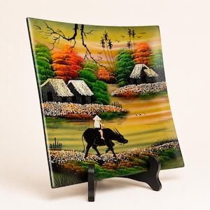 May include: A square decorative plate featuring a painted landscape of a rural river scene with thatched-roof huts, lush green and orange trees, and a person riding a black water buffalo in the foreground, displayed on a black plastic stand.