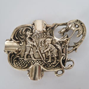 May include: A polished brass ashtray featuring an ornate relief design of two figures standing amidst scrolling foliage. The rectangular tray has rounded edges, three integrated cigarette rests, and a decorative handle shaped like a mythical creature.