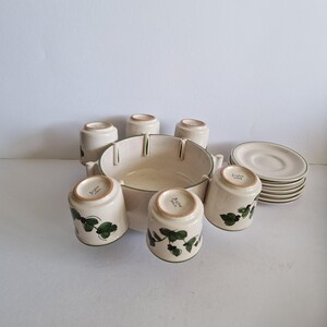Puede incluir: Un juego de vajilla de cerámica color crema, que incluye un cuenco grande con borde verde, tazas y platillos a juego. Las tazas presentan un diseño de hojas verdes. El juego incluye platillos apilados.