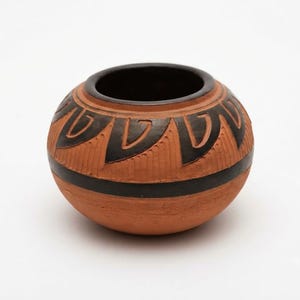 Puede incluir: Una vasija redonda de terracota con una abertura ancha y un interior marrón oscuro. El exterior presenta un acabado mate de color marrón rojizo con patrones geométricos negros en forma de pétalos y una banda horizontal negra.