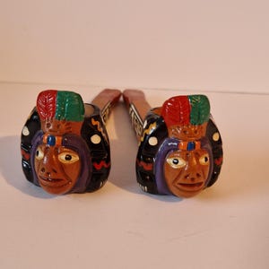 Puede incluir: Dos pipas decorativas de madera con caras de jefes nativos americanos. Las pipas tienen detalles en marrón, negro, rojo, verde y morado. Cada pipa tiene una cara tallada con un tocado de plumas.