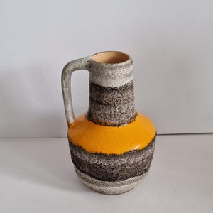 May include: A small, ceramic pitcher with a handle. The pitcher features horizontal bands of gray, brown, and orange. The interior is a light beige color. The handle is gray and the pitcher has a textured surface.