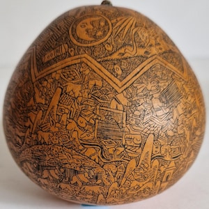 Può includere: Una zucca marrone chiaro con intricati disegni neri incisi. L'opera d'arte raffigura un paesaggio dettagliato con figure, edifici e animali. La zucca ha una forma arrotondata e una piccola sommità scura.