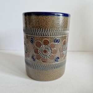 Pode incluir: Um vaso cilíndrico de cerâmica com uma superfície castanha salpicada e texturizada. O vaso apresenta uma borda azul escura e é decorado com flores castanhas em relevo, detalhes azuis e padrões geométricos. O design é vintage e decorativo.